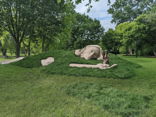A person sits beside a large, grassy sculpture of a reclining figure in a park surrounded by trees.