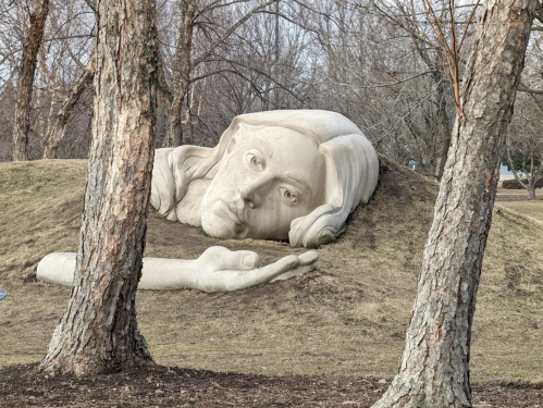 A large, sculpted face and hand emerging from a grassy hill, surrounded by bare trees in a park setting.