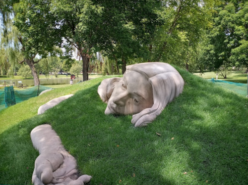 A large, abstract sculpture of a reclining figure partially covered by grass in a park setting.