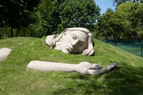 A large, sculpted head and arm emerging from a grassy hill, surrounded by trees under a clear blue sky.