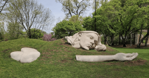 A large, abstract sculpture of a face and arm emerging from a grassy hill in a park, surrounded by trees.