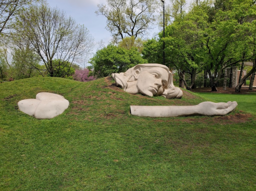 A large, abstract sculpture of a face and arm emerging from a grassy hill in a park, surrounded by trees.
