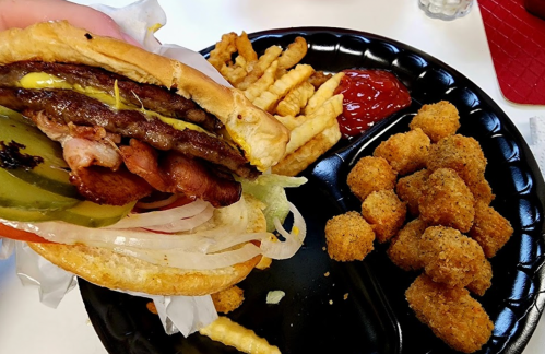 A hand holding a burger with bacon, pickles, and lettuce, alongside fries and tater tots on a black plate.