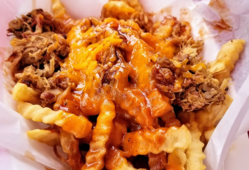 A close-up of loaded fries topped with pulled pork and melted cheese, served in a paper container.