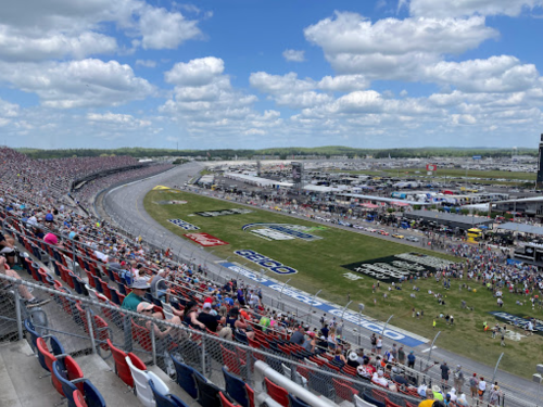 Aerial view of a crowded racetrack with fans in the stands and cars on the track under a partly cloudy sky.