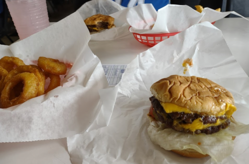 A cheeseburger with two patties and lettuce, served with a side of onion rings in a basket lined with paper.