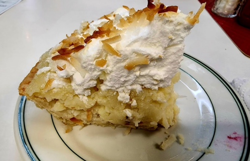 A slice of coconut cream pie topped with whipped cream and toasted coconut flakes on a white plate.
