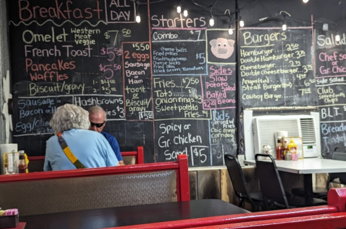 A diner menu on a chalkboard featuring breakfast, starters, burgers, and salads with prices, and two patrons seated.