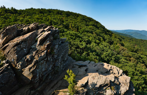 A rocky outcrop overlooks a lush green hillside under a clear blue sky.