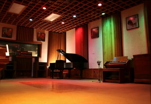A dimly lit recording studio featuring a grand piano, an organ, and colorful wall lights with framed portraits.