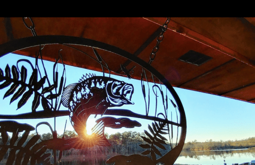 A metal fish sculpture hangs against a sunset over a calm lake, surrounded by trees.