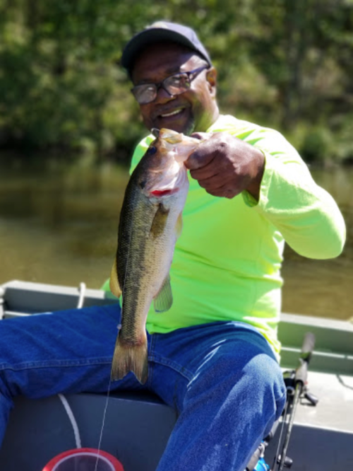 A smiling man in a bright green shirt holds a large bass while fishing from a boat on a calm lake.