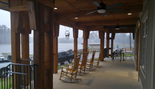 A covered porch with wooden rocking chairs overlooks a misty lake, surrounded by trees and parked cars.