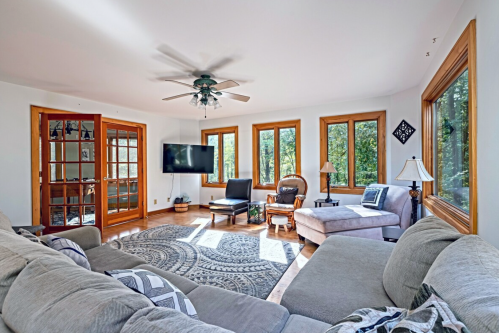 Bright living room with large windows, a cozy sofa, armchair, and a TV, featuring a patterned rug and wooden accents.