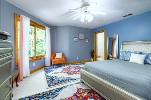 A cozy bedroom with a gray bed, colorful rug, and a window letting in natural light.