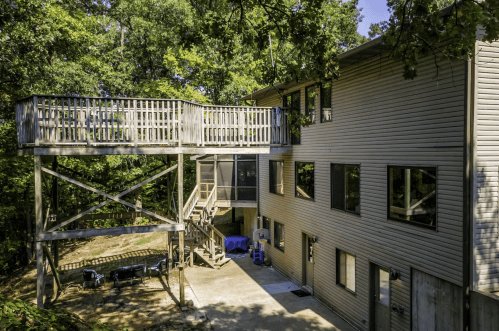 A two-story house surrounded by trees, featuring a large deck and outdoor stairs leading to the ground level.