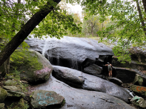 A person climbs a rocky surface near a waterfall, surrounded by trees and greenery.