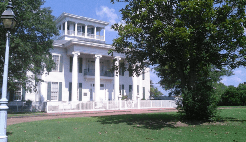 A large white historic house with columns, surrounded by greenery and a tree, under a clear blue sky.