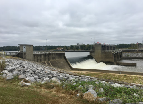A concrete dam with water flowing over it, surrounded by rocky terrain and cloudy skies.