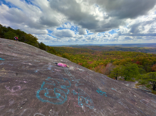 A rocky landscape with colorful graffiti, overlooking a valley of trees and clouds in the sky.