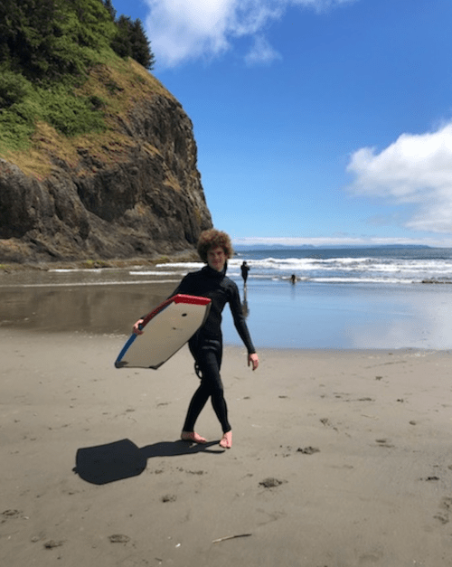 A person in a wetsuit walks along the beach holding a surfboard, with waves and cliffs in the background.