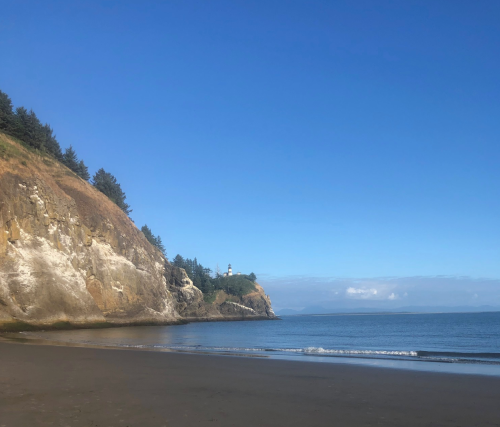 A serene beach scene with a rocky cliff, trees, and a lighthouse in the distance under a clear blue sky.