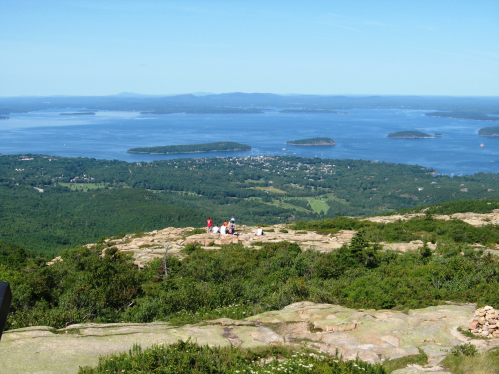 A scenic view from a mountain overlooking a lake with islands, surrounded by lush greenery and a clear blue sky.