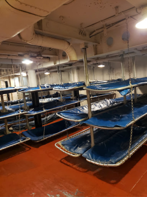 Interior of a ship's sleeping quarters with multiple metal bunk beds and blue mattresses, illuminated by overhead lights.