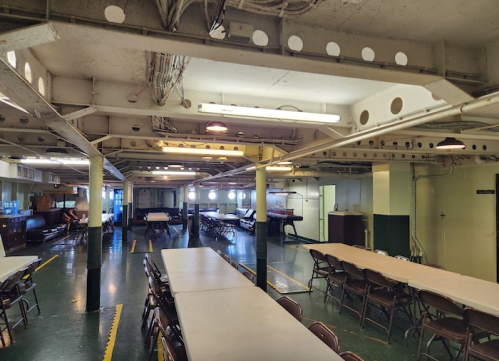 Interior of a spacious room with tables and chairs, featuring industrial-style beams and large windows.