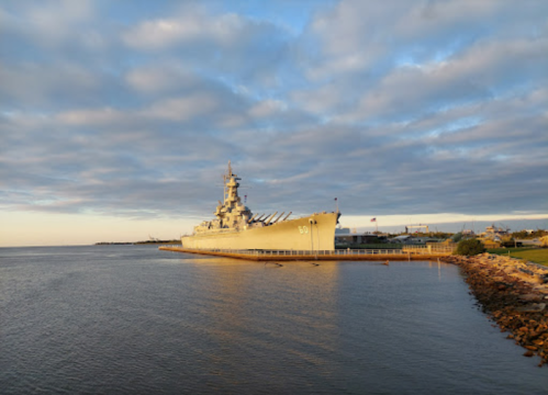 A battleship docked at a waterfront under a cloudy sky during sunset, with calm waters in the foreground.