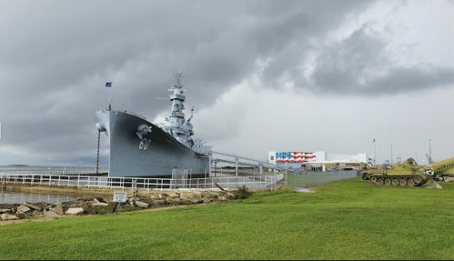 A battleship docked at a museum, with a tank displayed nearby and stormy clouds overhead.