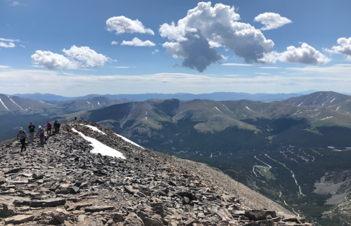 Hikers traverse a rocky mountain ridge with expansive views of valleys and distant peaks under a partly cloudy sky.