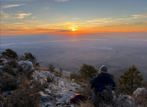 A person sits on a rocky ledge, watching a vibrant sunset over a vast landscape.