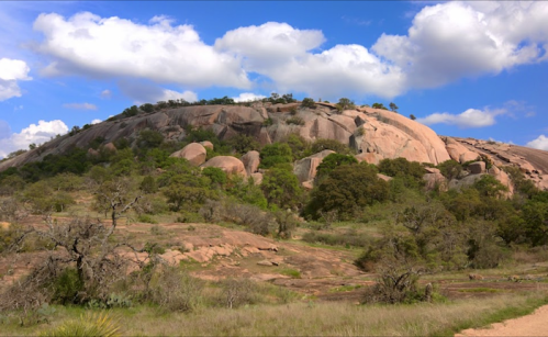 A rocky hill surrounded by greenery under a blue sky with fluffy clouds.