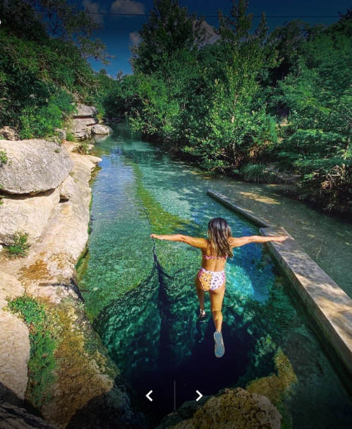 A person in a swimsuit jumps into a clear, blue river surrounded by lush greenery and rocky banks.