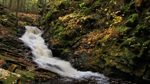 A serene waterfall cascades over rocky terrain, surrounded by lush greenery and autumn foliage.
