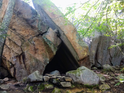 A large, triangular rock formation surrounded by trees and smaller stones in a natural setting.