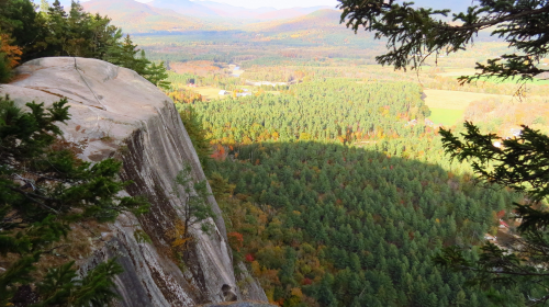 A panoramic view from a rocky cliff overlooking a dense forest and valley with autumn foliage.