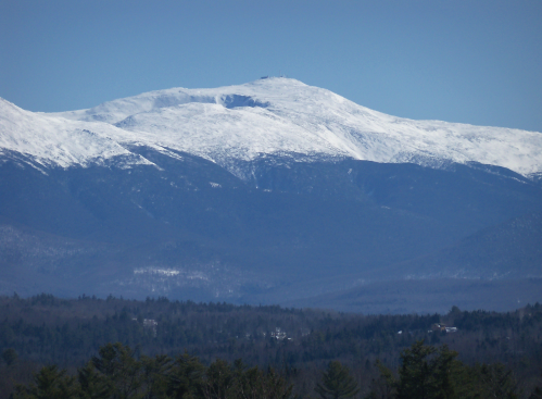 Snow-capped mountain peak under a clear blue sky, surrounded by a forested landscape.