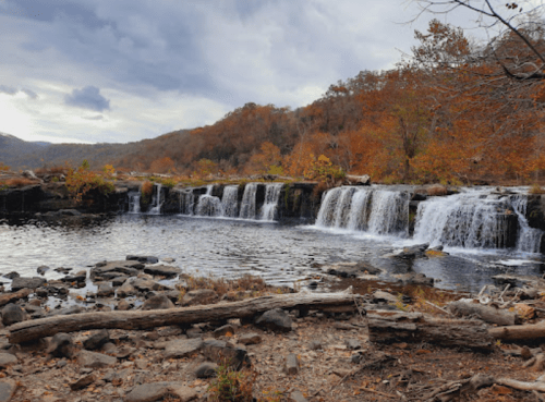 A serene waterfall cascades over rocks, surrounded by autumn foliage and a cloudy sky.