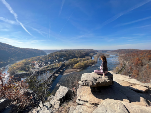 A person sits on a rock overlooking a river and autumn landscape under a clear blue sky.