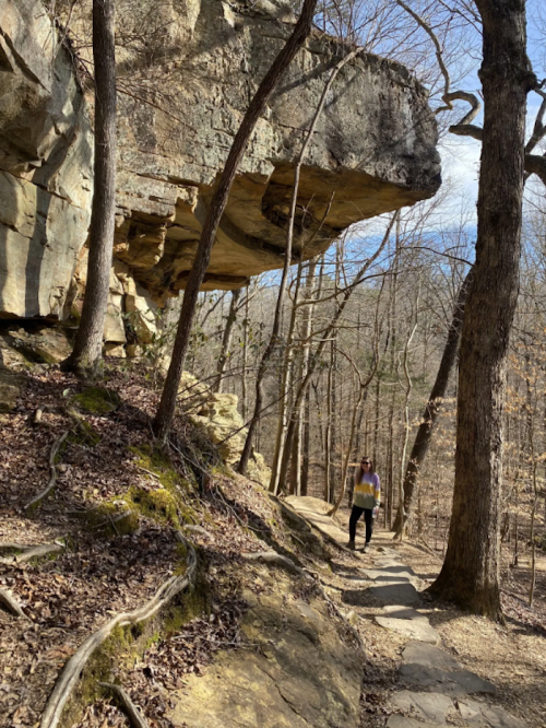 A person walks along a rocky trail surrounded by trees and a large overhanging rock formation.