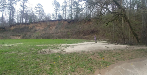A person stands on a sandy area near a grassy field, with a forested hillside in the background.