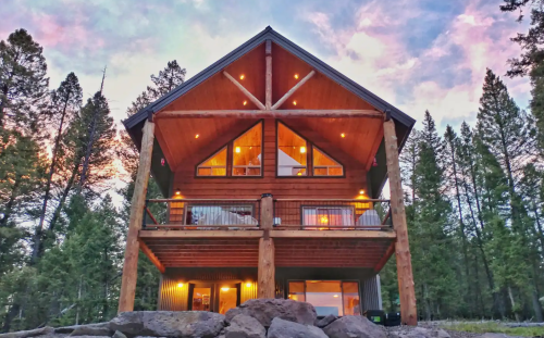 A wooden cabin with large windows and a balcony, surrounded by trees under a colorful sunset sky.