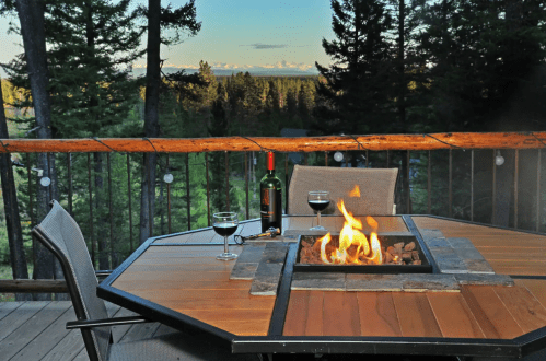 A cozy outdoor setting with a fire pit, wine glasses, and a bottle, surrounded by trees and mountains in the background.