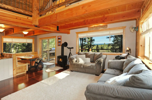 Cozy living room with wooden beams, large windows, a gray sofa, and a view of trees outside. Warm and inviting atmosphere.