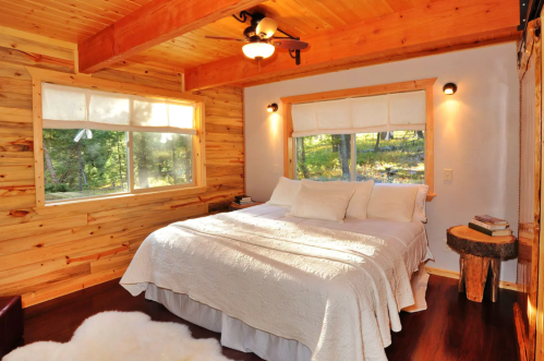 Cozy bedroom with wooden walls, a large bed, and windows overlooking a forested area. Soft lighting enhances the warmth.