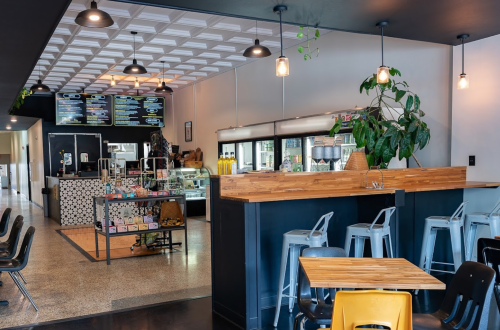 A modern café interior featuring a counter, seating, menu board, and plants, with a bright and inviting atmosphere.