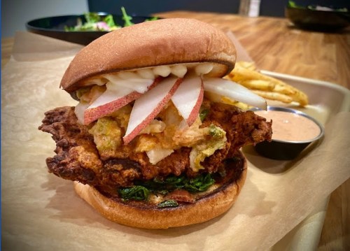 A crispy fried chicken sandwich topped with sliced pears and greens, served with a side of fries and dipping sauce.