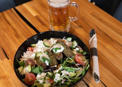 A bowl of salad with greens, tomatoes, cucumbers, and falafel, alongside a glass of beer on a wooden table.
